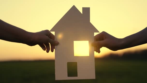 Símbolo de casa y felicidad. Feliz concepto de construcción de viviendas para la familia. La familia sostiene la casa de papel al atardecer, el sol brilla a través de la ventana. Sueño con construir, comprar casa para niños — Vídeos de Stock