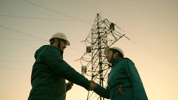 Trabalho em equipe e engenheiros, parceiros apertando as mãos, bom trabalho. Conceito de negócio. Mulher e homem trabalham juntos. Trabalhadores de energia em capacetes brancos de proteção estão verificando linha de energia on-line usando tablet computador — Fotografia de Stock