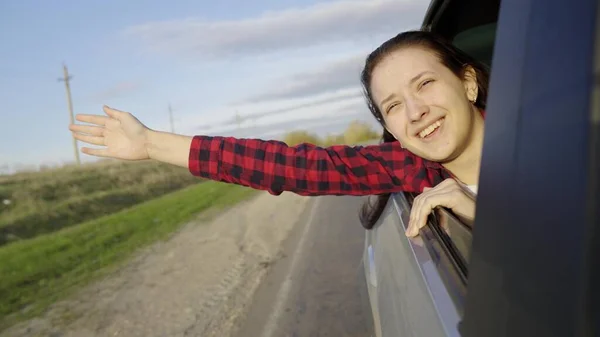 Ragazza libera viaggia in auto cattura il vento con la mano dal finestrino dell'auto. Giovane donna con i capelli lunghi siede nel sedile posteriore della macchina, si allunga verso il finestrino e cattura bagliore di sole. Un adolescente viaggia in macchina. — Foto Stock