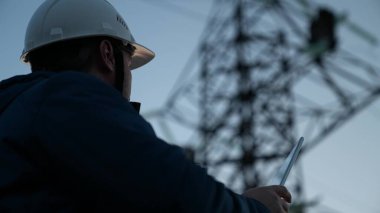 Koruyucu beyaz miğferdeki güç mühendisi, güç hattını kontrol etmek için bilgisayar tableti, enerji sistemine uzaktan erişim kullanarak kontrol ediyor. Gün batımında yüksek voltajlı elektrik hatları. temiz enerji