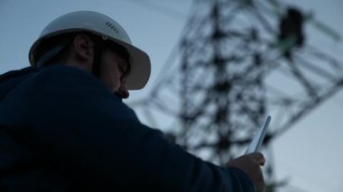 Koruyucu beyaz miğferdeki güç mühendisi, güç hattını kontrol etmek için bilgisayar tableti, enerji sistemine uzaktan erişim kullanarak kontrol ediyor. Gün batımında yüksek voltajlı elektrik hatları. temiz enerji