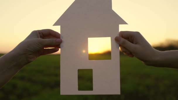 La familia sostiene la casa de papel al atardecer, el sol brilla a través de la ventana. Movimiento de cámara a ventana. Hogar y símbolo de felicidad. Concepto de construcción de casa para la familia. Mi sueño es construir, comprar casa para niños. — Vídeos de Stock