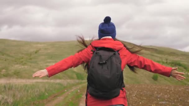 A free, happy traveler runs into mountains with open arms, wind blows her hair. Young woman travels with backpack enjoying beautiful scenery of mountains and hills. Travel and adventure concept — Stock Video