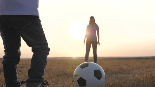 Happy family and child have fun playing a soccer ball in park. The kid kicks ball. Mom and daughter play football on field in rays of the sunset. Teamwork of the child and mom. Healthy sports family