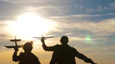 Uçakta oynayan çocukların silueti. Çocuklar gökyüzünün ve güneşin önünde ellerinde bir uçakla. Uçma hayalleri. Mutlu çocukluk kavramı. İki kız gün batımında oyuncak bir uçakla oynuyorlar..
