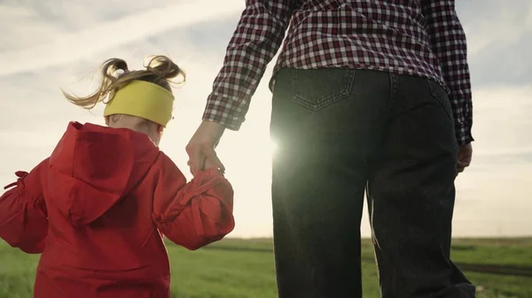 Feliz niño de la familia y mamá están caminando en el parque tomados de la mano. La pequeña hija sostiene a sus madres de la mano. Hija despreocupada y mamá caminan juntas a través del campo. Concepto de una familia infantil saludable. — Foto de Stock