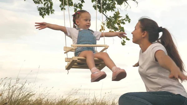 Mãe de família feliz e filhinha estão brincando no parque de verão. Uma mãe está balançando sua filhinha alegre em um balanço debaixo de uma árvore. Uma mãe brinca com o filho. Conceito de infância feliz. — Fotografia de Stock