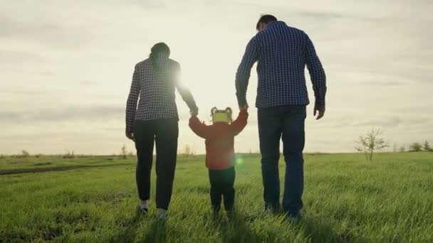 Fröhliche Familienwanderung im Park im Frühling bei Sonnenuntergang. Mama, Papa und Tochter gehen, Händchen haltend, Kind hüpft auf dem grünen Gras. Glückliche gesunde Kindheit. Familienwochenende — Stockvideo