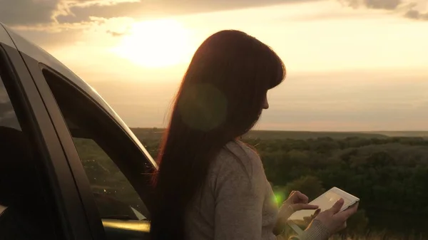 Turysta ze smartfonem zatrzymał się na kempingu samochodem, patrząc na zachód słońca z góry. Turysta kierowca trzyma nowoczesny gadżet w rękach, odpoczynek. Wolna kobieta, podróżująca samochodem, sprawdzająca trasę w nawigatorze. — Zdjęcie stockowe