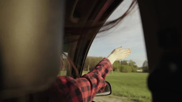 Wolna kobieta prowadzi samochód, łapie wiatr przez okno samochodu. Dziewczyna z długimi włosami siedzi na przednim siedzeniu samochodu, wyciąga rękę przez otwarte okno, cieszyć się podróżą — Zdjęcie stockowe