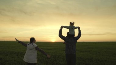 Mutlu aile, çocuk ve ebeveynler gün batımı siluetinde parkta birlikte koşuyorlar. Baba, kız, anne uçak gibi koşuyor, pilot olma ve uçma hayalleri kuruyor. Neşeli aile ve küçük çocuk tarlada oynuyorlar.