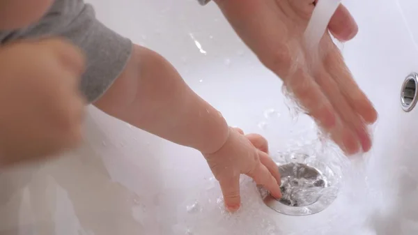 The kid learns to wash his hands by himself. A small child washes his hands with his mother over the sink. Mother helps her child to wash hands at home before breakfast. Play with your child in the — Stock Photo, Image