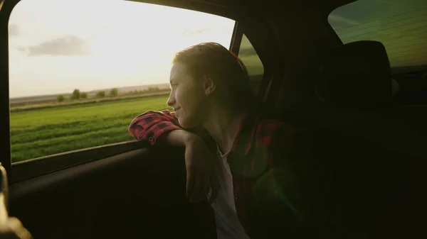 Wolna kobieta podróżuje samochodem chwyta wiatr ręką z okna samochodu. Dziewczyna z długimi włosami siedzi na przednim siedzeniu samochodu, wyciągając rękę przez okno i chwytając blask zachodzącego słońca — Zdjęcie stockowe