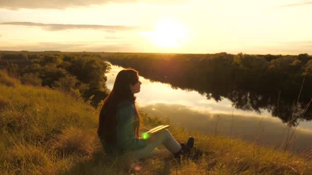 La joven viajera lee el libro en un alto. Chica turista con un libro en la montaña al sol sobre el río. Un estudiante lee un libro de texto en el parque al aire libre en los rayos del atardecer. Oración en la naturaleza. — Vídeos de Stock