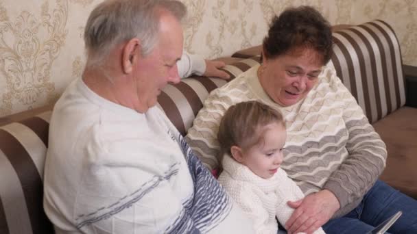 Famille heureuse, grands-parents modernes jouent avec un enfant, une fille assise avec une tablette en ligne à la maison. Enfant, fille montre quelque chose en tablette aux grands-parents. Couple âgé avec petite-fille et gadget — Video