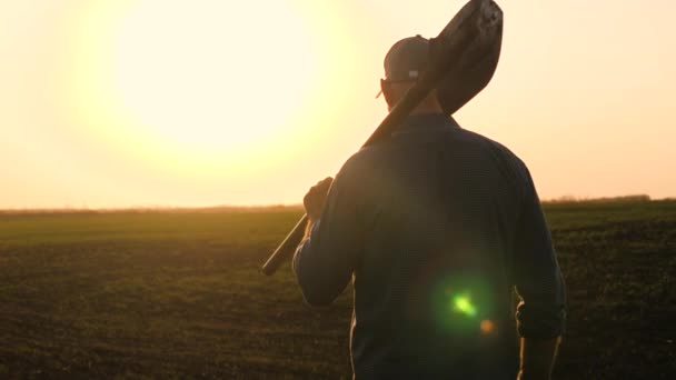 A farmer with a shovel in his hands walks across the field with a shovel in the sun. An agronomist walks on a black fertile plowed land at sunset. Worker with a shovel in a field at sunset. — Stock Video