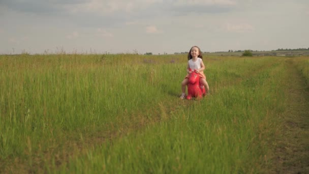 O miúdo joga em campo com o seu cavalo de brinquedo favorito. Menina pequena feliz está jogando no parque e saltando em um burro inflável do brinquedo. Menina, filha a brincar no campo ao ar livre. Família e infância. — Vídeo de Stock