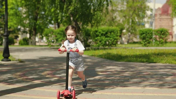 Uma menina aprende a andar de scooter. A criança feliz está a brincar no parque. Uma criança saudável anda de scooter pela cidade na rua. O conceito de uma infância feliz, família, saúde. Família fim de semana ao ar livre. — Fotografia de Stock