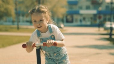 Küçük bir kız scooter sürmeyi öğreniyor. Mutlu çocuk parkta oynuyor. Sağlıklı bir çocuk şehirde scooter kullanıyor. Mutlu bir çocukluk, aile, sağlık kavramı. Aile hafta sonu dışarıda..