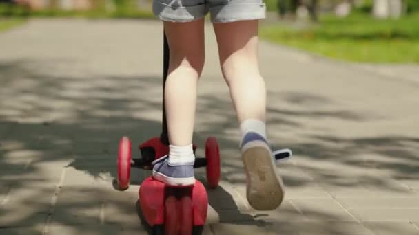 Seorang gadis kecil belajar mengendarai skuter. Keluarga akhir pekan di luar. Anak yang bahagia sedang bermain di taman. Seorang anak yang sehat mengendarai skuter di sekitar kota di jalan. Konsep masa kecil yang bahagia, keluarga, kesehatan. — Stok Video