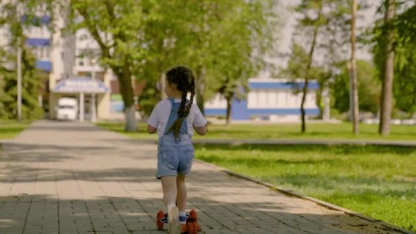 Een gezond kind rijdt op een scooter door de stad op straat. Een klein meisje leert op een scooter rijden. Gelukkig kind speelt in het park. Het concept van een gelukkige jeugd, familie, gezondheid. Familie weekend buiten. — Stockvideo