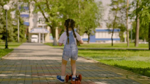 Een gezond kind rijdt op een scooter door de stad op straat. Een klein meisje leert op een scooter rijden. Gelukkig kind speelt in het park. Het concept van een gelukkige jeugd, familie, gezondheid. Familie weekend buiten. — Stockfoto