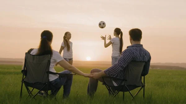Happy family. Mom and dad are sitting in tourist chairs in field, children are playing ball in sun. Travelers, mom, dad, children on vacation play together at sunset in park. Healthy family, childhood