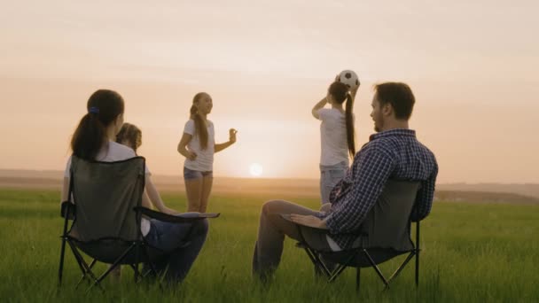 Lycklig familj. Mamma och pappa sitter i turiststolar på fältet, barnen leker boll i solen. Resenärer, mamma, pappa, barn på semester leker tillsammans vid solnedgången i parken. Friska familjen, barndom — Stockvideo