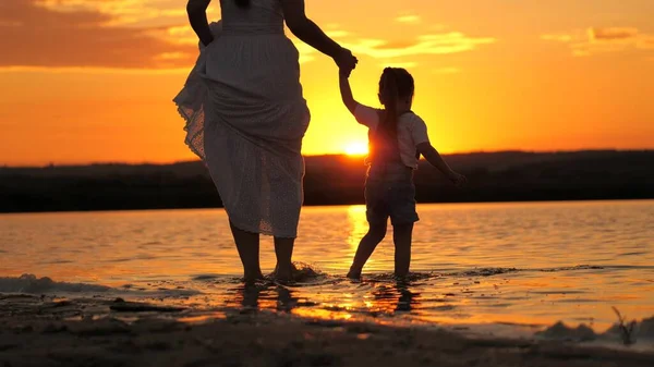 Silueta de una mujer y un niño en la playa al atardecer. Feliz familia, mamá camina con el niño descalzo en el agua, uniendo las manos en los rayos de una hermosa puesta de sol. Madre junto con su hija en la playa. — Foto de Stock