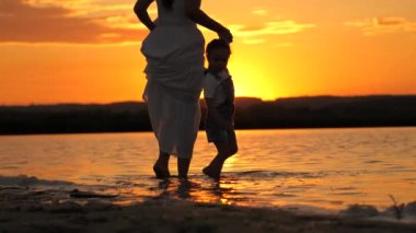 Gün batımında sahilde bir kadın ve bir çocuğun silueti. Mutlu bir aile, anne suda çıplak ayakla yürür, güzel bir gün batımında el ele tutuşur. Anne ve kızı kumsalda..