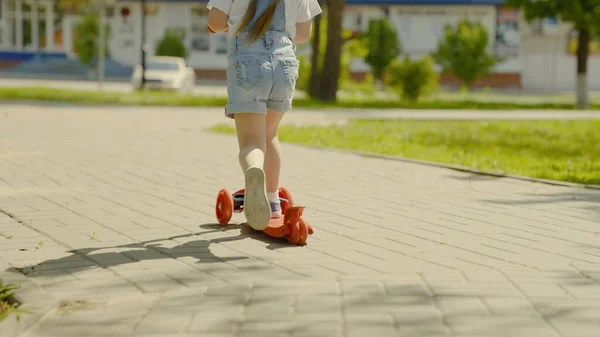 Un niño sano monta un scooter por la ciudad en la calle. El niño feliz está jugando en el parque. Una niña aprende a montar en scooter. Fin de semana familiar al aire libre. El concepto de una infancia feliz, familia, salud. —  Fotos de Stock