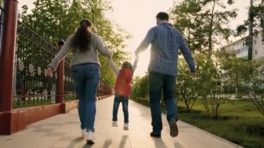 Mutlu aile sokakta parkta koşuyor, bahar ve yaz aylarında el ele tutuşuyorlar. Mutlu sağlıklı bir çocukluk. Takım çalışması. Anne, baba, kız şehir parkında oynuyorlar, mutlu çocuk ebeveynlerin elini tutuyor ve zıplıyor..