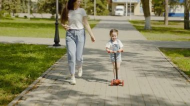 Mutlu bebek ve anne parkta oynuyorlar. Küçük kız scooter sürmeyi öğreniyor. Doğada aile hafta sonu. Sağlıklı çocuk şehir caddesi boyunca annesinin gözetiminde scooter kullanıyor. Aile, çocukluk