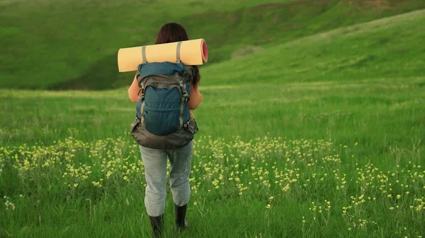 Uma jovem caucasiana ativa com mochila vai em caminhada. Menina Viajante viaja na grama verde e flores, meditação, ecoturismo, caminhadas. Mulher livre viajante caminhando pelo prado no verão. — Fotografia de Stock