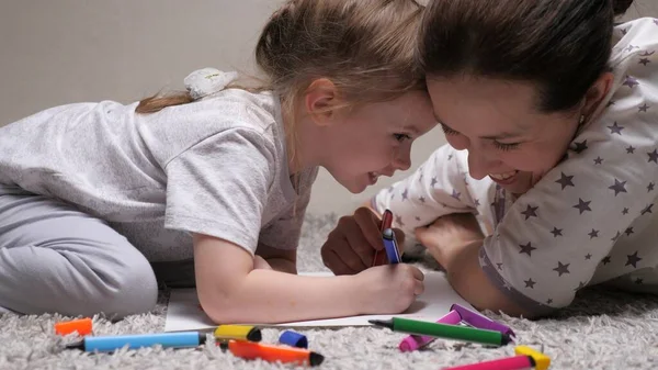 Child and Mom, nanny, teaches girl to draw. Happy family playing together at home laughing. A mother helps her daughter learn to draw on paper, coloring with multi-colored pencils and felt-tip pens. — Stock Photo, Image