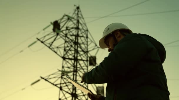 Power Engineer en casque blanc de protection vérifie la ligne électrique en ligne à l'aide d'une tablette informatique, accès à distance au système énergétique, pour le contrôle et la sécurité. Lignes électriques à haute tension au coucher du soleil. énergie propre — Video