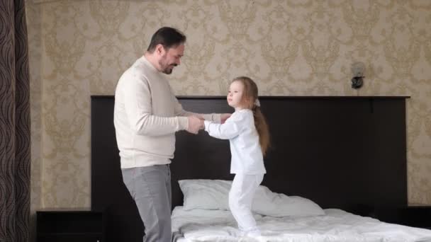 Щаслива родинна дитина, дочка і тато, тримаються за руки, стрибають у ліжко. Батько весело сміється, граючи активну гру зі своєю милою донькою, дитиною в спальні. Танцює вранці. Тато вихідний — стокове відео