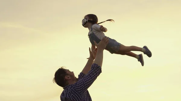 Silhouet van vader en kind, kleine meisjes, spelen, genieten van de zonsondergang in het park in de natuur op zomerdag. Gelukkig gezin, vader speelt met kind als piloot, gooit hem in de lucht, kind verheugt zich en lacht. — Stockfoto