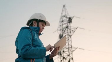 Beyaz kasklı bir kadın güç mühendisi tabletteki elektrik sensörlerinden elde edilen verileri kullanarak güç hattını kontrol ediyor. Gün batımında yüksek voltajlı elektrik hatları. Dağıtım ve elektrik tedariği. temiz enerji