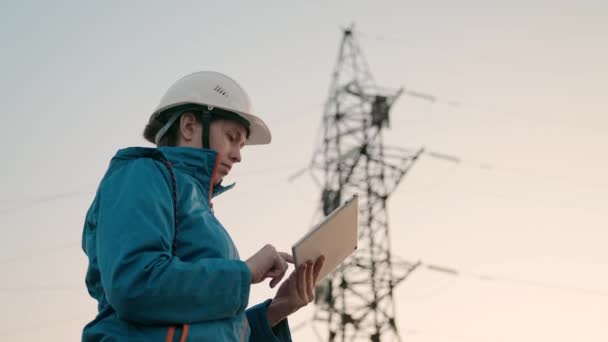 Een vrouwelijke power engineer in witte helm inspecteert de stroomleiding met behulp van gegevens van elektrische sensoren op een tablet. Hoogspanningsleidingen bij zonsondergang. Distributie en levering van elektriciteit. schone energie — Stockvideo