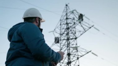 Koruyucu beyaz miğferdeki güç mühendisi, güç hattını kontrol etmek için bilgisayar tableti, enerji sistemine uzaktan erişim kullanarak kontrol ediyor. Gün batımında yüksek voltajlı elektrik hatları. temiz enerji