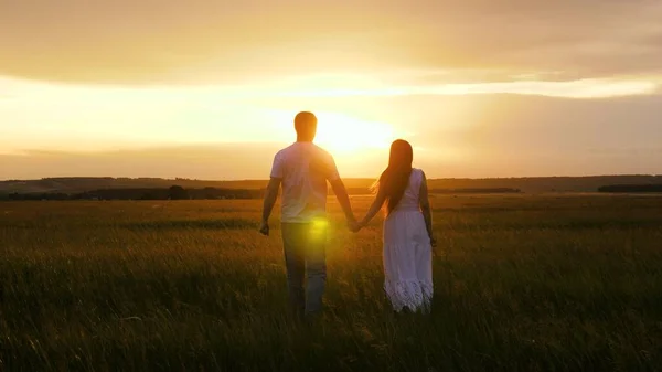 Silhouet van man en vrouw bij zonsondergang. Romantisch afspraakje en liefde in de natuur. Een mooi meisje en een jongen lopen hand in hand in het veld, in de zon op het groene gras. Een verliefd jong stel reist.. — Stockfoto