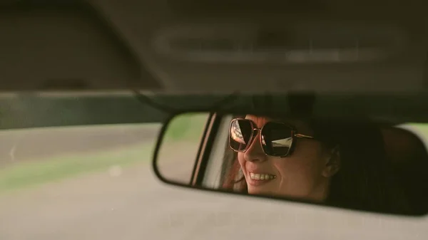 Piękna młoda kobieta podróżująca samochodem, uśmiechnięta, radująca się. Młoda kobieta w okularach przeciwsłonecznych w lusterku wstecznym. Kierowca prowadzi samochód. Weekendy, samochód, przygoda, droga i sport. — Zdjęcie stockowe