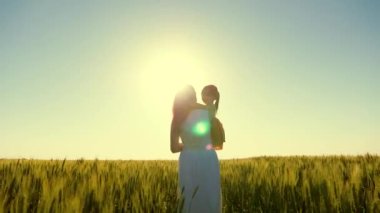 Anne ve çocuk buğday tarlasında yürüyorlar. Bebek annesinin kollarında. Anne, çocuk, kız yeşil buğday tarlasında yürüyor, sarılıyor ve öpüşüyorlar. Mutlu aile gezileri. Çiftçi kadın ve çocuk tarlada