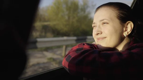 Uma menina livre viaja de carro, olha pela janela do carro e sonha, dirigindo. Uma jovem passageira senta-se no banco de trás de um carro, aproveitando a viagem e pegando o brilho do sol. — Fotografia de Stock