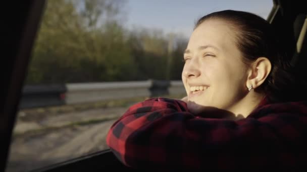 Una chica libre viaja en coche, mira por la ventana del coche y sueña, conducir. Una joven pasajera se sienta en el asiento trasero de un coche, disfrutando del viaje y cogiendo el resplandor del sol. — Vídeos de Stock