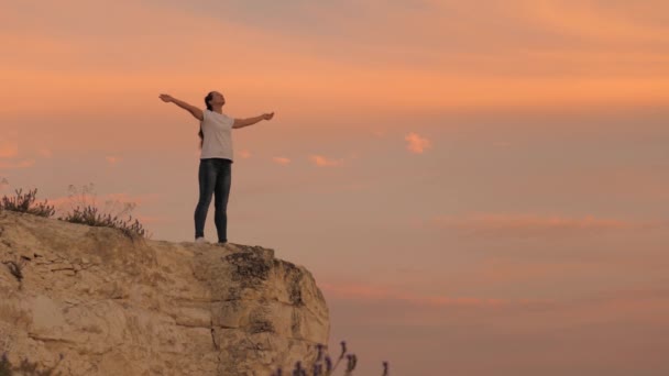 Girl hiker relaxes and meditate on top of hill. Young happy woman raises her hands up, standing on top of a mountain above sea against backdrop of blue sky. Watching sunset with a beautiful landscape. — Stock Video