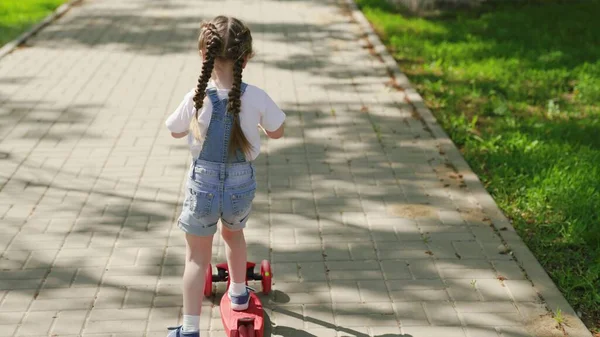 Маленькая девочка учится ездить на скутере. Семейные выходные на открытом воздухе. Счастливый ребенок играет в парке. Здоровый ребенок ездит на скутере по городу на улице. Концепция счастливого детства, семьи, здоровья. — стоковое фото