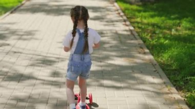 Küçük bir kız scooter sürmeyi öğreniyor. Aile hafta sonu dışarıda. Mutlu çocuk parkta oynuyor. Sağlıklı bir çocuk şehirde scooter kullanıyor. Mutlu bir çocukluk, aile, sağlık kavramı..
