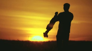 Mutlu bir aile, baba çocukla oynuyor, çocuğu kollarında döndürüyor, yazın güneşe karşı neşeli ve kahkahalar atıyor. Baba ve çocuğun silueti, doğada günbatımının tadını çıkaran küçük kızlar.